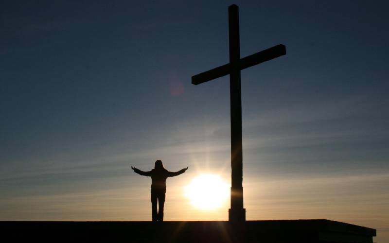 A person standing in front of a cross at sunset.