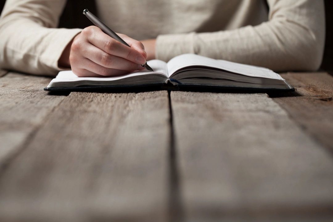 A person writing in a notebook on top of a wooden table.